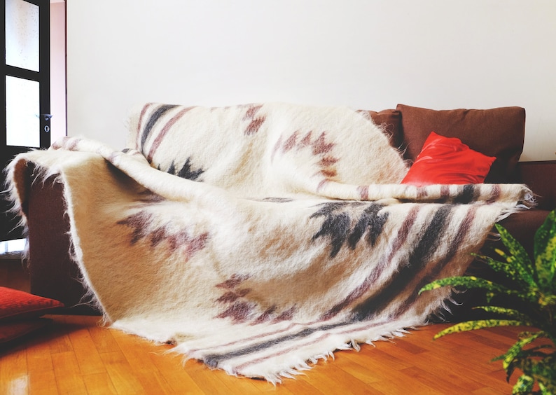 May include: A white, brown, and gray patterned throw blanket draped over a brown couch with a red pillow. The blanket has a fringe border.
