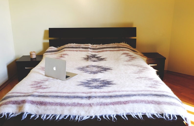 May include: A white blanket with a geometric pattern in shades of gray and pink is spread across a bed with a dark wood headboard. A silver laptop computer is open and resting on the blanket.