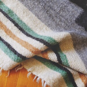 May include: A woven throw blanket with a white background and stripes of green, black, and brown. The blanket is folded and has a frayed edge.