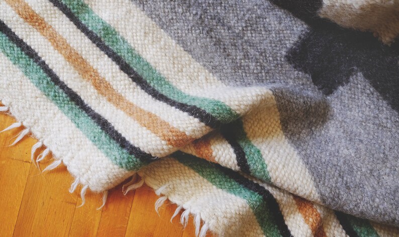 May include: A woven throw blanket with a white background and stripes of green, black, and brown. The blanket is folded and has a frayed edge.