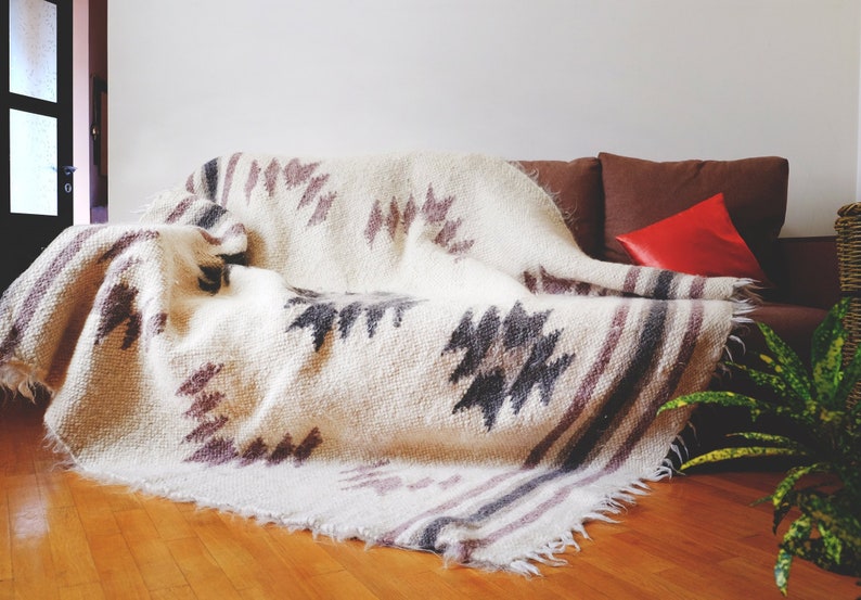 May include: A white throw blanket with a geometric pattern in shades of gray and purple. The blanket is draped over a brown couch with a red pillow.