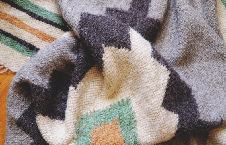 May include: A close-up of a woven rug with a geometric pattern in shades of gray, white, green, and orange. The rug is made of wool and has a soft, textured surface.