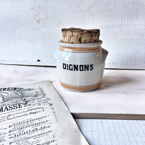 Peut inclure: Un pot à oignons en céramique blanche avec un bouchon en liège et le mot "OIGNONS" en noir. Le pot a une bande beige au milieu et est posé sur une surface blanche à côté d'une partition ancienne.