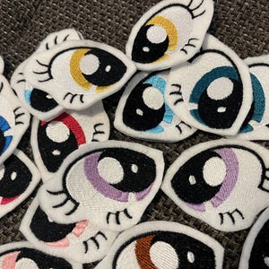 Könnte beinhalten: Eine Sammlung weißer Filz-Augen-Patches mit gestickten Details. Die Augen sind schwarz mit verschiedenen Farbakzenten, darunter Blau, Gelb, Lila und Braun.