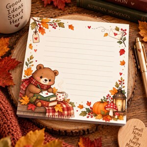 Könnte beinhalten: Ein quadratischer Notizblock mit linierten Seiten und einem herbstlichen Rand. Das Design zeigt einen braunen Bären mit rotem Schal, einen Kürbis, eine Laterne, Pilze sowie bunte Herbstblätter und kleine Herzmotive auf dem Papier.