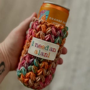 May include: A handmade, colorful crochet can cozy. The cozy is a mix of pink, orange, green, and brown yarn. A small white tag with the embroidered text "I need an alani" is attached. An orange can of "Orange Kiss" is inside the cozy.