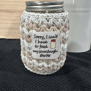 May include: A white and beige crocheted jar cozy with a white embroidered patch that says "Sorry, I can't I have to feed my sourdough starter".