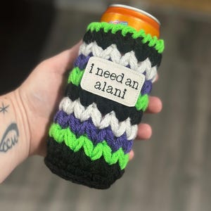 May include: A handmade crochet can cozy in black, white, purple, and green chevron pattern. The cozy has a small white tag that reads "i need an alani". The cozy is designed to fit a standard size can.