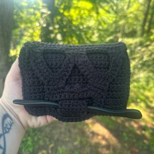 May include: A black crocheted hair accessory featuring a rectangular base with a textured, raised geometric pattern. A smooth, solid black hair stick is inserted horizontally through a central loop on the front of the piece to secure it in place.