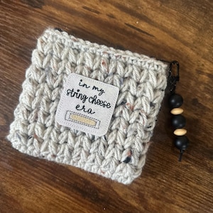May include: A crocheted, speckled gray pouch with a small white label that reads "in my string cheese era." The pouch has a black zipper and a black beaded charm with wooden accents.