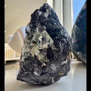Puede incluir: Una formación rocosa rugosa, de color gris oscuro, con grupos de cristales translúcidos de color verde claro incrustados. Los cristales tienen formas geométricas afiladas, que contrastan con la textura rugosa de la roca. La roca está sobre una superficie blanca.