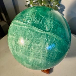 Op de afbeelding: Een grote, gepolijste groene bol met een natuurlijk, getextureerd oppervlak. De bol heeft een levendige groene tint, met lichtere strepen en een subtiele glans. Hij staat op een kleine, vierkante houten basis, die de bolvorm benadrukt. Het is een decoratief item.