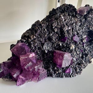 May include: A large, rough-textured rock formation with clusters of vibrant purple fluorite crystals. The dark matrix contrasts with the bright, geometric crystal structures, creating a striking mineral specimen.