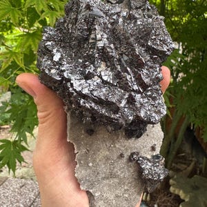May include: A large, dark, metallic mineral specimen featuring a cluster of sharp, angular, crystalline structures with a reflective, gunmetal-gray surface, attached to a flat, light-gray, textured, rocky matrix base with smaller crystal deposits.