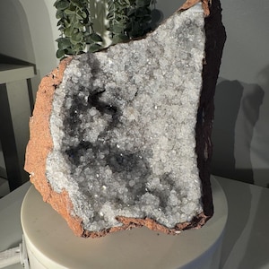 Op de afbeelding: Een grote, ruwe geode met een roodbruine buitenkant en een sprankelend, kristallijn interieur. De kristallen zijn voornamelijk wit en grijs en vullen de holte van de rots. De geode wordt gepresenteerd op een witte, cirkelvormige standaard.