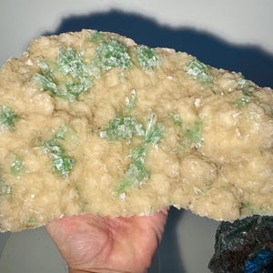 May include: A large, irregularly shaped mineral specimen with a creamy beige base and clusters of bright green crystals. The crystals are scattered across the surface, creating a textured appearance. The mineral is held in a hand.