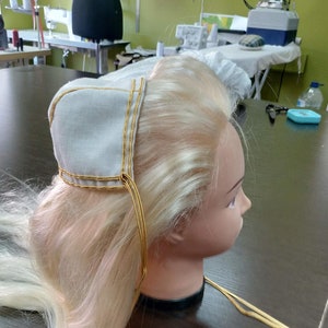 May include: A white fabric head covering with gold trim and long ties. The head covering is on a mannequin with long blonde hair.