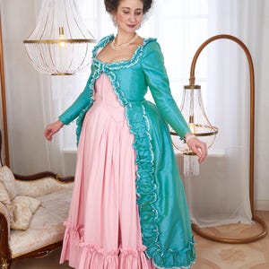May include: A pink and teal historical gown. The dress features a pink bodice and skirt with ruffled trim. A teal jacket with long sleeves and white trim is worn over the dress. The gown is reminiscent of 18th-century fashion.