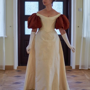 May include: A woman wearing a white gown with brown puffed sleeves. The gown has a fitted bodice and a full skirt. She is standing in a room with a wooden floor.
