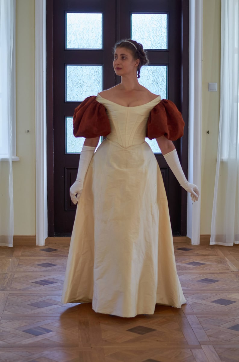 May include: A woman wearing a white gown with brown puffed sleeves. The gown has a fitted bodice and a full skirt. She is standing in a room with a wooden floor.