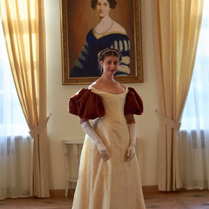 May include: A woman in a white gown with brown puffed sleeves stands in a room with gold curtains and a portrait of a woman in a blue dress. The woman in the portrait is wearing a pearl necklace.