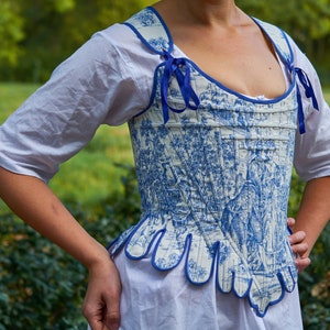 May include: A white and blue patterned corset with blue ribbon ties. The corset has a scalloped bottom edge.