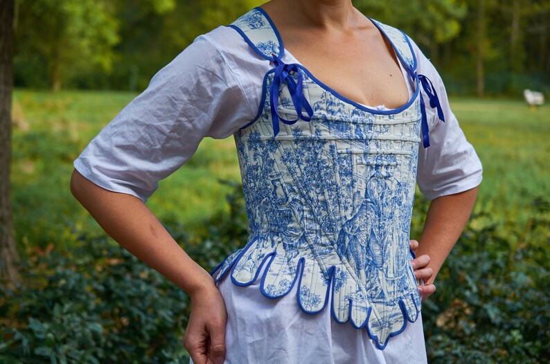 May include: A white and blue patterned corset with blue ribbon ties. The corset has a scalloped bottom edge.