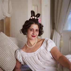 May include: A woman wearing a white empire-waist dress and a pearl necklace. Her hair is styled with decorative braids and a pink flower. She is reclining on a bed with a striped pillow, creating a vintage aesthetic.
