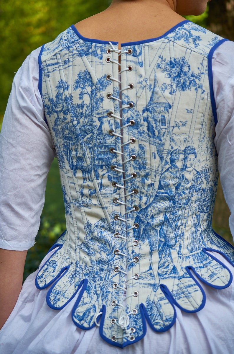 May include: A white and blue patterned corset with a blue trim and a lace-up back. The corset has a scalloped bottom edge.
