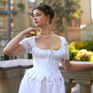 May include: A woman in a white, ruffled, corset-style top with a pearl necklace. She is standing on a balcony with a building in the background.