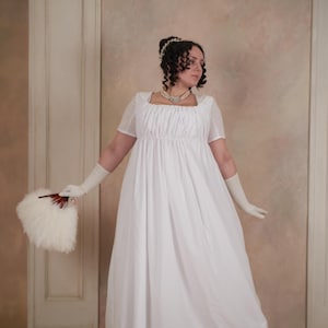 May include: A white Regency-style dress with short sleeves and a square neckline. The dress features a gathered bodice and a flowing skirt. The person is holding a white feathered fan and wearing long white gloves. The background is a neutral color.