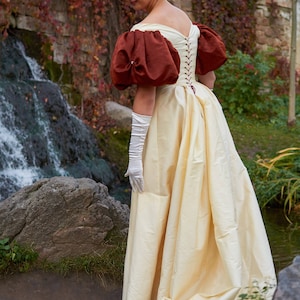 May include: A woman wearing a white gown with a brown bodice and puffed sleeves. The gown is cinched at the waist with a red ribbon. She is wearing white gloves and a pearl headband. The woman is standing in front of a waterfall and a stone wall.