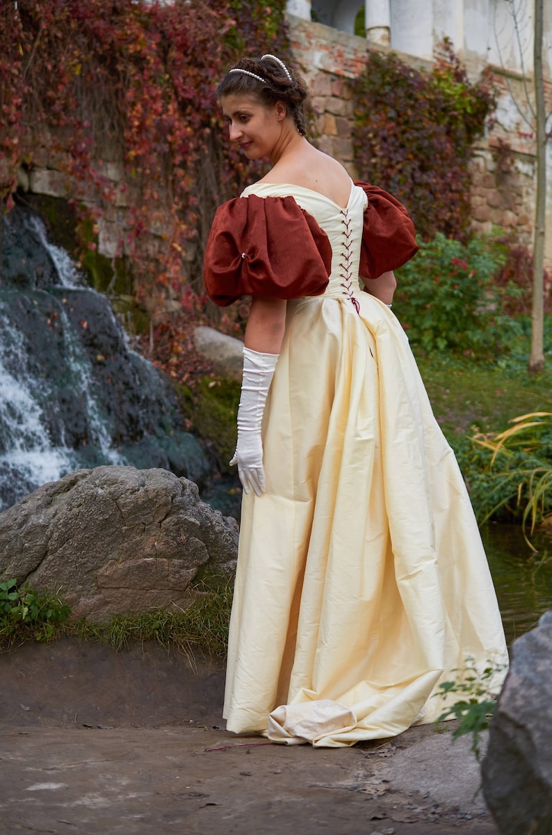 May include: A woman wearing a white gown with a brown bodice and puffed sleeves. The gown is cinched at the waist with a red ribbon. She is wearing white gloves and a pearl headband. The woman is standing in front of a waterfall and a stone wall.