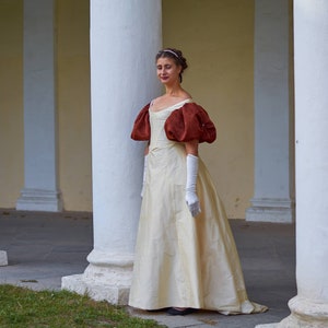 May include: A woman in a white gown with large, puffed, burgundy sleeves stands in a white columned hallway. She is wearing white gloves and a headband.