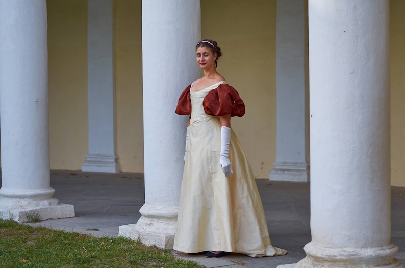 May include: A woman in a white gown with large, puffed, burgundy sleeves stands in a white columned hallway. She is wearing white gloves and a headband.