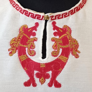 May include: White garment with red and gold embroidery. The neckline features a red geometric pattern. The front has a symmetrical design of two stylized creatures in red with gold accents. A small gold button secures the neckline.