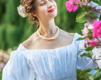 May include: A woman in a white dress with puffed sleeves and a pearl necklace is standing in a garden and smelling pink roses.