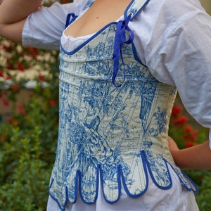 May include: A white and blue patterned corset with a blue ribbon tie. The corset has a scalloped bottom edge and is made of a lightweight fabric.