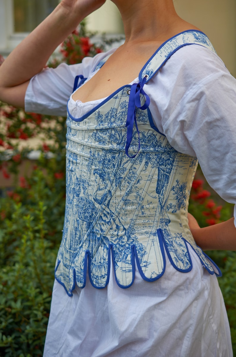 May include: A white and blue patterned corset with a blue ribbon tie. The corset has a scalloped bottom edge and is made of a lightweight fabric.