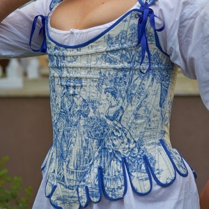 May include: A white and blue patterned corset with a blue ribbon detail. The corset has a scalloped bottom edge and is made of a lightweight fabric.