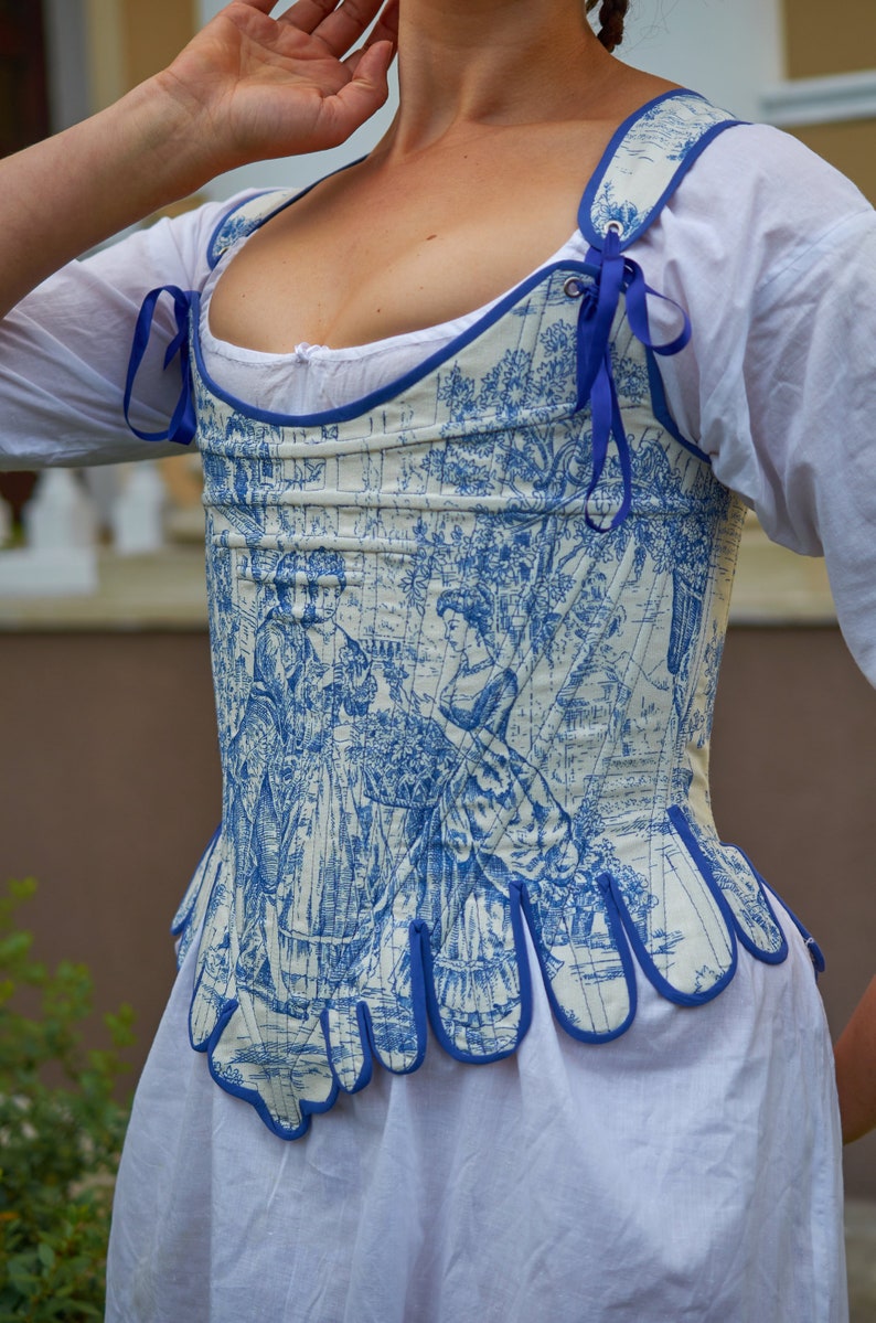 May include: A white and blue patterned corset with a blue ribbon detail. The corset has a scalloped bottom edge and is made of a lightweight fabric.