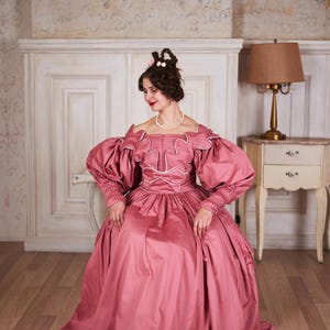 May include: A woman in a pink, floor-length gown with puffed sleeves and white trim sits on a white chair in a room with a white wainscoting and a lamp on a side table.