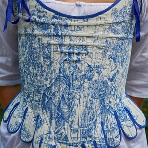 May include: A white and blue corset with a blue trim and a floral pattern. The corset has a scalloped bottom edge and is tied with blue ribbons.
