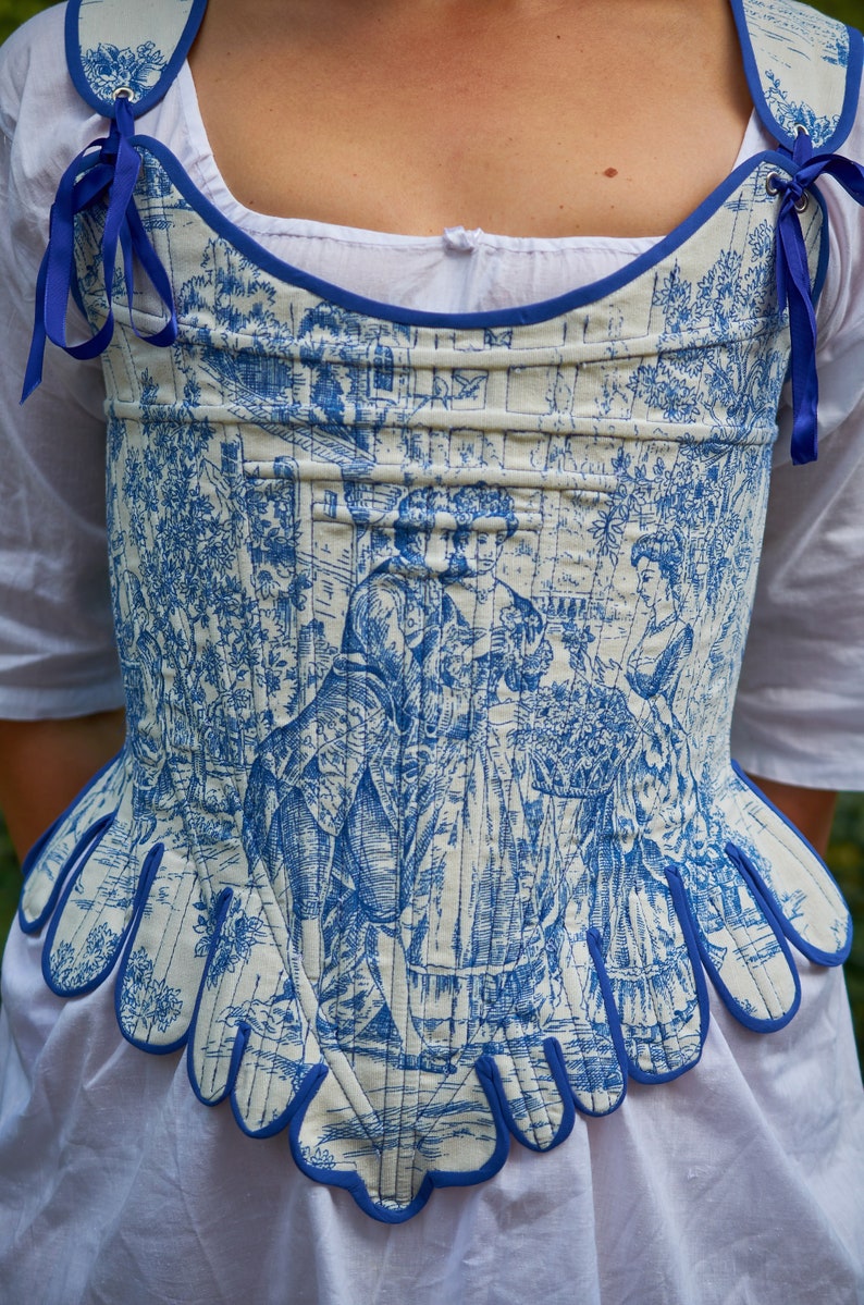 May include: A white and blue corset with a blue trim and a floral pattern. The corset has a scalloped bottom edge and is tied with blue ribbons.
