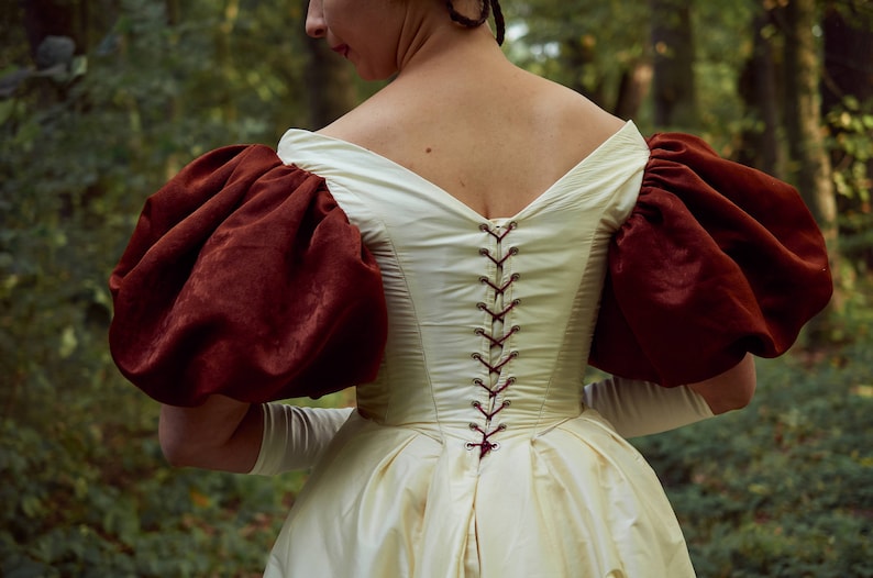 May include: A white dress with a deep V-neckline and a lace-up back. The dress has large, puffed sleeves made of a burgundy fabric.