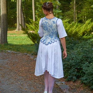 May include: A woman wearing a white dress with a blue and white patterned bodice. The bodice has a scalloped edge and is cinched at the waist. She is standing on a path in a forest.