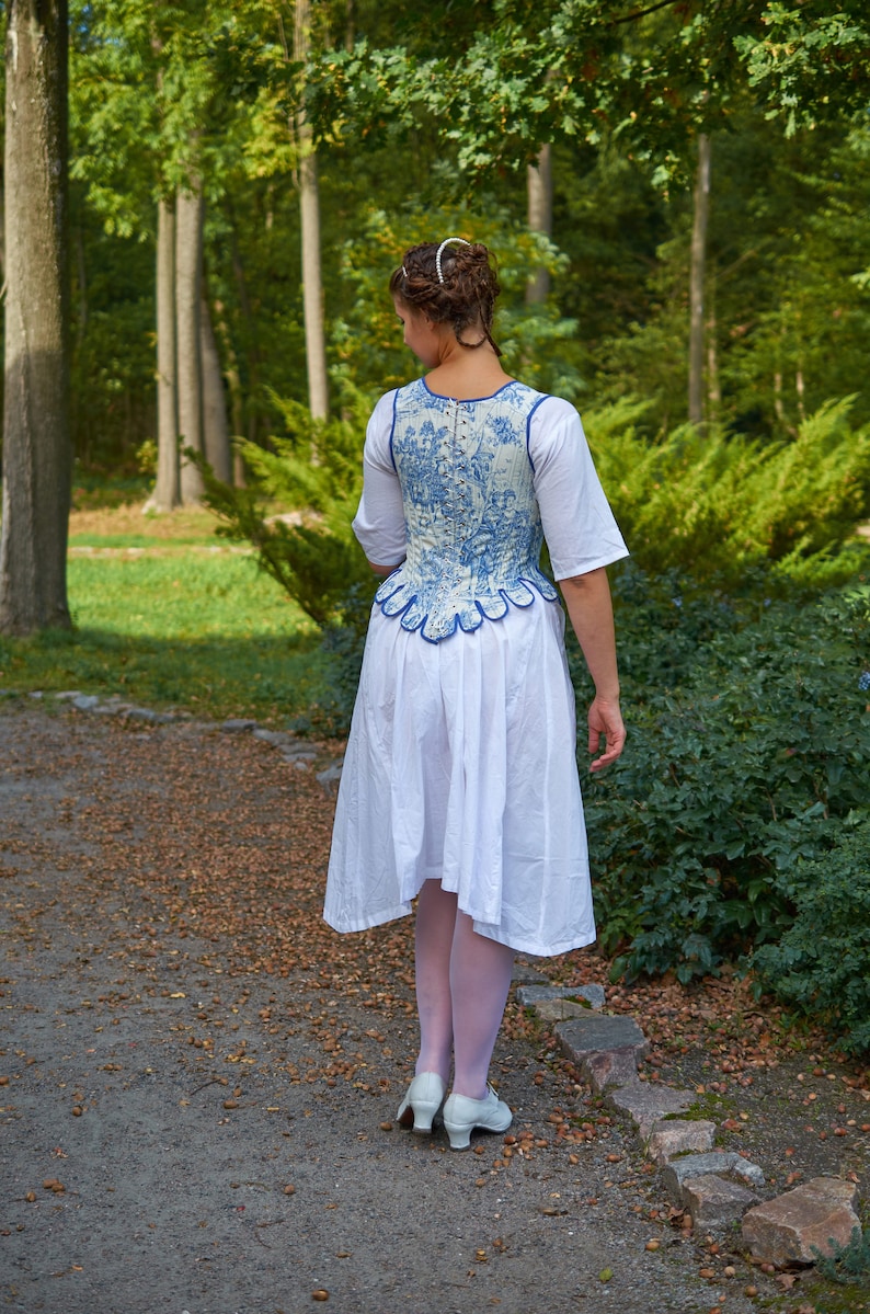 May include: A woman wearing a white dress with a blue and white patterned bodice. The bodice has a scalloped edge and is cinched at the waist. She is standing on a path in a forest.