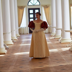 May include: A woman in a long, cream-colored gown with maroon puffed sleeves stands in a hallway with white columns. The gown has a fitted bodice and a full skirt. She is wearing white gloves and has her hands clasped in front of her.