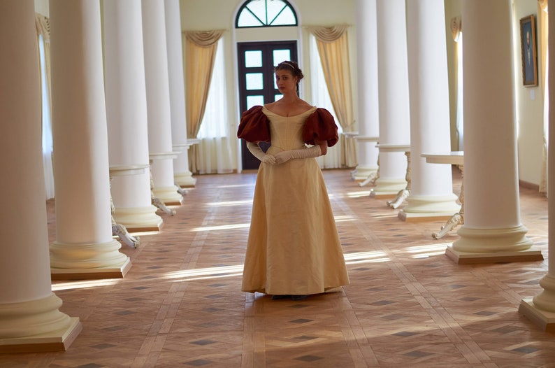 May include: A woman in a long, cream-colored gown with maroon puffed sleeves stands in a hallway with white columns. The gown has a fitted bodice and a full skirt. She is wearing white gloves and has her hands clasped in front of her.