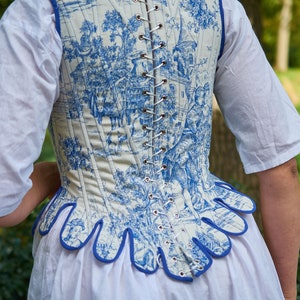 May include: A white corset with a blue and white toile pattern. The corset has a blue trim and is laced up the back.